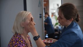 Close-up view of make-up artist applying cream makeup foundation using brush on adult blond woman's face in beauty salon. Soft focus. Real time handheld video. Make-up and beauty industry theme.