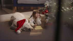 Sweet small caucasian boy laying on floor with big dog in beautifully decorated room crafting letter to Santa Claus. Cinematic ad. Boy squints with pleasure in anticipation of receiving treasured gift