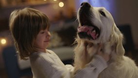 Close up boy and his white dog. Boy pulls dog by neck, happy dog breathes with his mouth open as if smiling, settled down by Christmas tree. Joyous occasion of New Year is near. Cinematic advertising