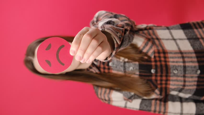 Vertical footage of serious young brunette woman 25s holding unsatisfied sad face emoticon, posing isolated over plain red color background wall in studio. Concept angry, stressed, negative emotion | Shutterstock HD Video #1111670685