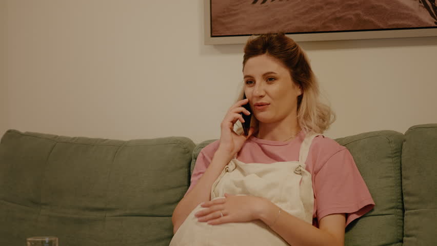 Pregnant female caressing her belly while talking on phone relaxing on couch. Future mom talking to the doctor having no health issues. Pregnancy and technology concept. | Shutterstock HD Video #1111680827