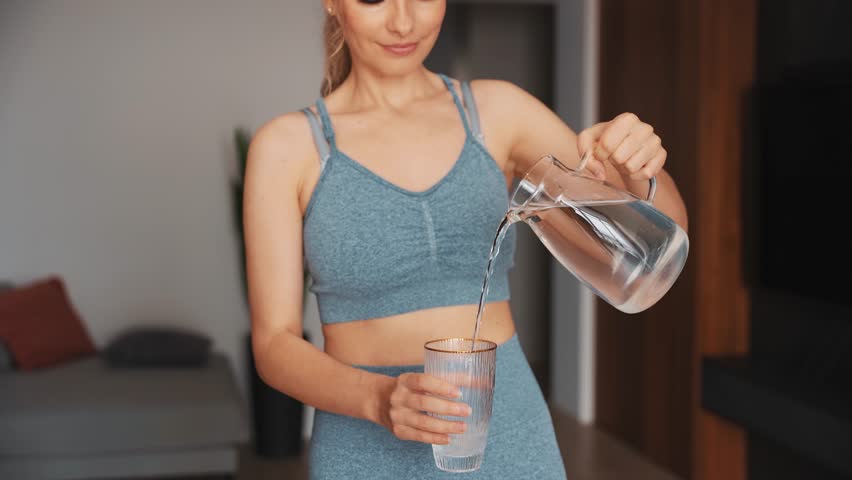 Joy young happy woman smiling face holds glass decanter in hands pours water into glass, drinks fresh cool clean water after hard workout sports at home. Girl in living room. sport fit yoga fitness | Shutterstock HD Video #1111683091