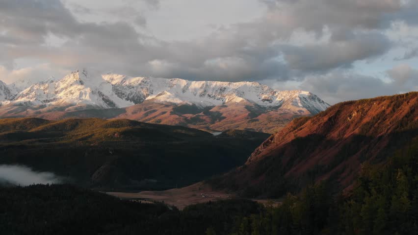Mountainous landscape, hilly terrain. Sun illuminates hills with gentle golden sunset rays, emphasizing splendor of natural beauty. Clouds bending over snow-capped mountain in distance. Aerial view
