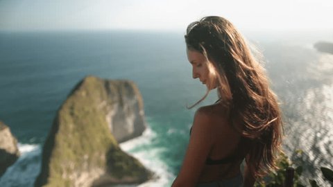 Slow motion shot girl stand on cliff, wind blowing long hair. Tourist woman enjoy aerial sea view landscape on Nusa Penida Island, Kelingking Secret Point Beach. Outdoor travel summer holiday vacation, videoclip de stoc