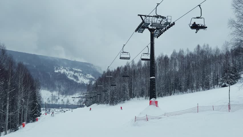 A chairlift at a ski resort on a cloudy winter day in the Ural Mountains. A lift on the background of factory pipes. Climbing the mountain. Gubakha, Perm Krai, 4K