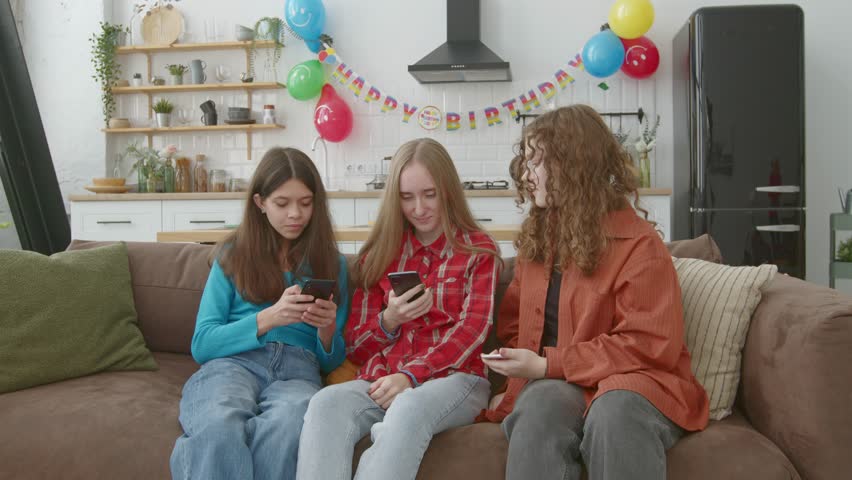 Positive lovely adolescent female friends sitting on sofa, networking and discussing social media content online using cellphones while enjoying leisure and preparing for birthday party at home.