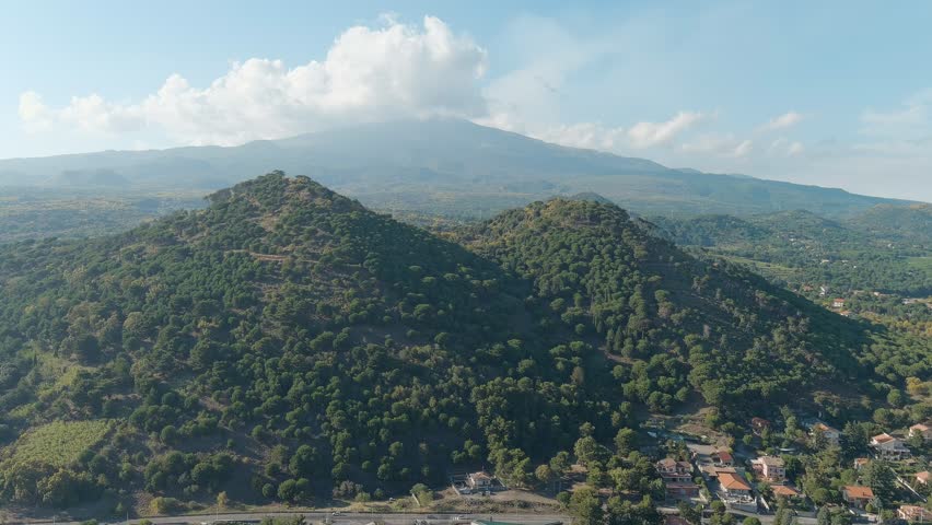 Nicolosi, Sicily, Italy. The Crateri Silvestri, two extinct volcanic cones formed during the 1892 eruption, rise from the southern slopes of Mount Etna blanketed in dense Mediterranean forest. An aerial perspective captures the rounded profiles of these pyroclastic cones, their slopes thick with pine and oak woodland that has reclaimed the ancient lava rock over more than a century. The massive silhouette of Mount Etna dominates the background, its summit partially veiled by drifting clouds against a bright blue sky. Below the forested cones, scattered rural buildings and a winding road mark the edge of human settlement. The scene is bathed in warm summer daylight, with rich greens contrasting against the darker volcanic earth and the hazy blue-gray mass of the active stratovolcano beyond. The slow forward drone movement reveals the layered volcanic terrain and the dramatic scale of Europe