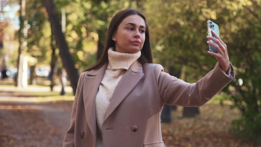 Young beauty woman walks in coat in autumn park, holds phone in hand and takes picture of herself on front camera and takes selfie or talks and communicates on video call. Leisure in nature landscape.
