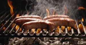 Flames seep through grill grate. Sausages are fried on hot grill. Cook lovingly turns sausages until they taste perfect. Fire coals give these delicious summer treats best flavor. Copy space, close-up