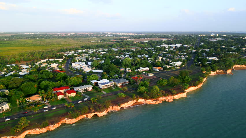 Aerial pull back of a small coastal town revealing cityscape in the distance. Coastline drone shot at sunset with blue water and city views
