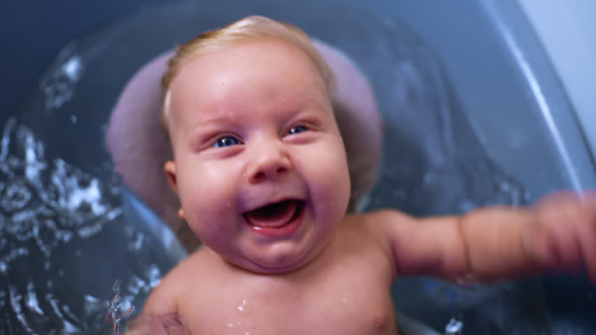 Sweet blue-eyed baby boy with fair hair lies in the wash tub. Happy kid waving his hands and feet while having a bath. Close up.