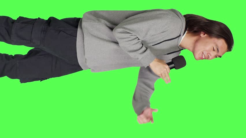 Side view of cool person doing karaoke session on camera, using microphone to sing modern songs and lyrics. Young adult having fun over greenscreen backdrop, creating music and singing tunes.