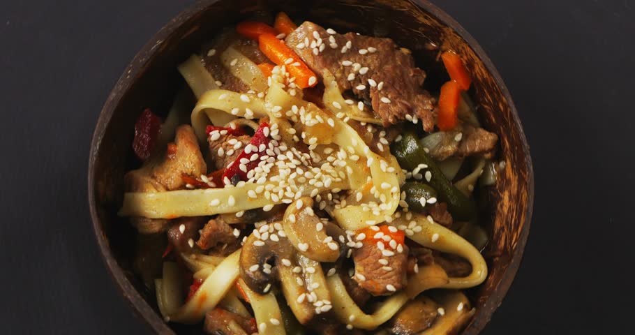 Wok udon with beef in a coconut shell plate sprinkled with sesame seeds in sauce rotates in a circle on a black background | Shutterstock HD Video #1111719671