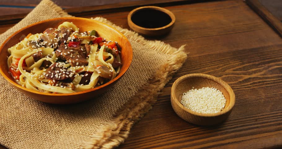 Wok udon with beef in a wooden plate on a salad sprinkled with sesame seeds rotates in a circle on a wooden table next to sesame seeds and soy sauce. | Shutterstock HD Video #1111719673