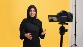 Smiling Muslim woman in black hijab talking shooting video use camera posing isolated on orange studio. Beautiful oriental young female blogger vlogger influencer recording media content interview