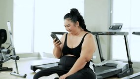 Young chubby Asian female wearing sportswear sitting in gym using smartphone technology search training course workout in social media to start exercising, obese woman change, break bad eating habits