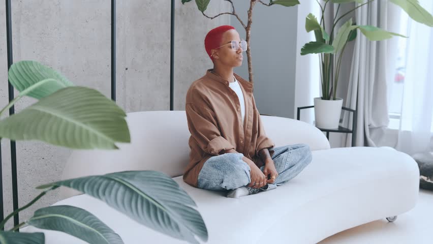 Young relaxed African American woman meditates sits on sofa in lotus position and practicing zen or yoga to achieve spiritual harmony and internal balance located in apartment. Millennial lifestyle | Shutterstock HD Video #1111740901