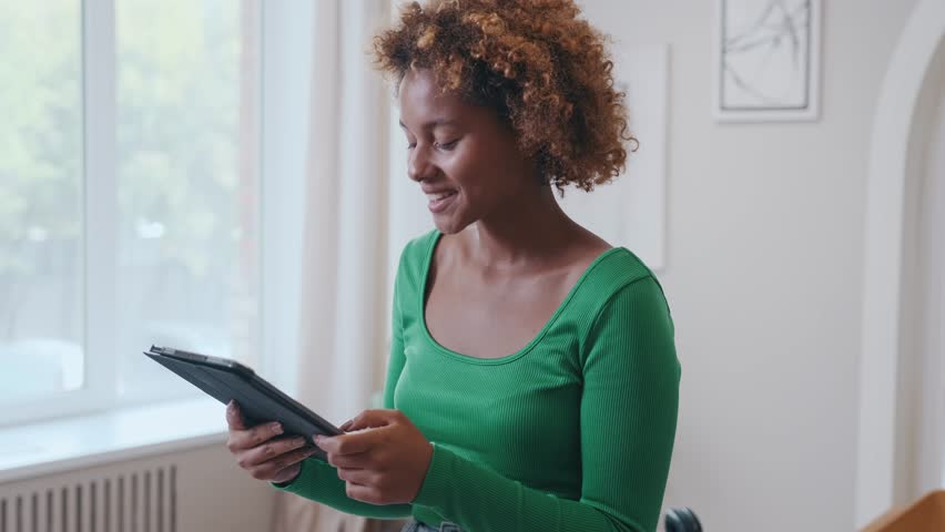 Young attractive happy African American woman freelancer with tablet computer in hands with smile performs online work and enjoys creating internet career stands in spacious apartment. | Shutterstock HD Video #1111740913