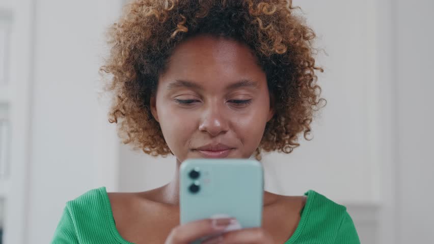 Young smiling cheerful African American woman enjoys using mobile phone and chatting with friends through online messengers or social networks and dating applications sits on sofa in own home. | Shutterstock HD Video #1111740915