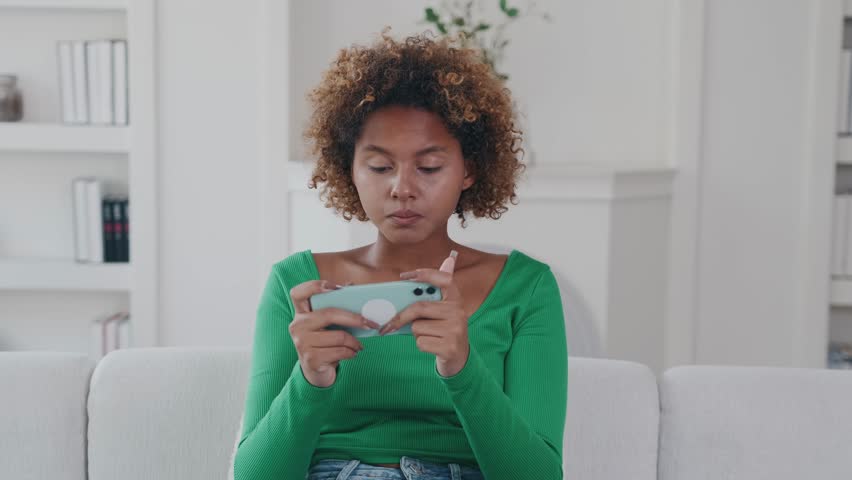 Young enthusiastic upset African American woman plays video games on mobile phone and loses mood after losing or accidentally making mistake sits on sofa in living room of apartment. | Shutterstock HD Video #1111740917