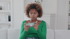 Young enthusiastic upset African American woman plays video games on mobile phone and loses mood after losing or accidentally making mistake sits on sofa in living room of apartment.
