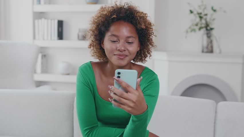 Young cheerful beautiful African American woman with phone in hands laughs watching stand-up show or comedy series with cool jokes that improve mood sits on sofa in living room with bookcases. | Shutterstock HD Video #1111740921