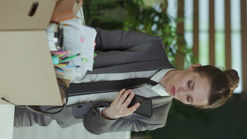 New job. stressed modern woman worker in modern green office in grey business suit with personal belongings in cardboard box speaking on a smartphone. Royalty-Free Stock Footage #1111741285