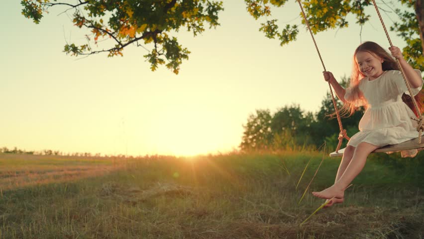 Child girl plays on wooden swing, dream fly. Happy little girl swings on swing in park under tree, sunset nature. Baby swing, kid girl smile in flight. Family happiness, dream, entertainment Concept
