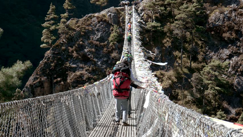 Trekking to Everest Base Camp. Tourists with backpacks walk on Hillary suspension bridge through gorge of Himalayan mountains. Prayer flags and ribbons are fluttering in wind. Rear view group hikers Royalty-Free Stock Footage #1111746705