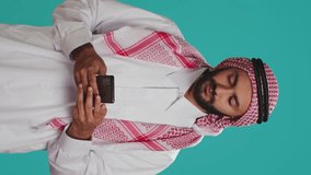 Vertical video Islamic guy in robe texting on phone while posing with confidence over blue background. Middle eastern person dressed in thobe and headscarf, sending online messages on smartphone app.
