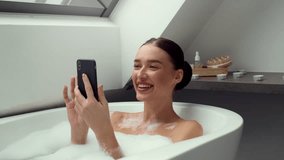 4K video footage, happy young woman, immersed in a foamy bath, looking at her smartphone, slow motion. The scene portrays a blend of relaxation and the modern digital connection in leisure moments.
