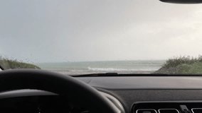 It is raining heavily, sea view is visible from inside the modern car, windshield wiper clean rain drop on windscreen, close-up