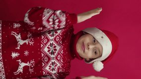 a cheerful Caucasian boy in a sweater and a Santa hat dances and looks at the camera. vertical video. studio shooting. concept holiday mood, christmas celebration