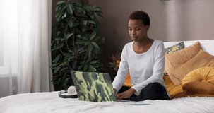 Exhausted tired african american dark skinned woman with shoty fashionable haircut sitting in lotos pose on bed wearing casual outfit working online from home freelancer concept. Female taking break.