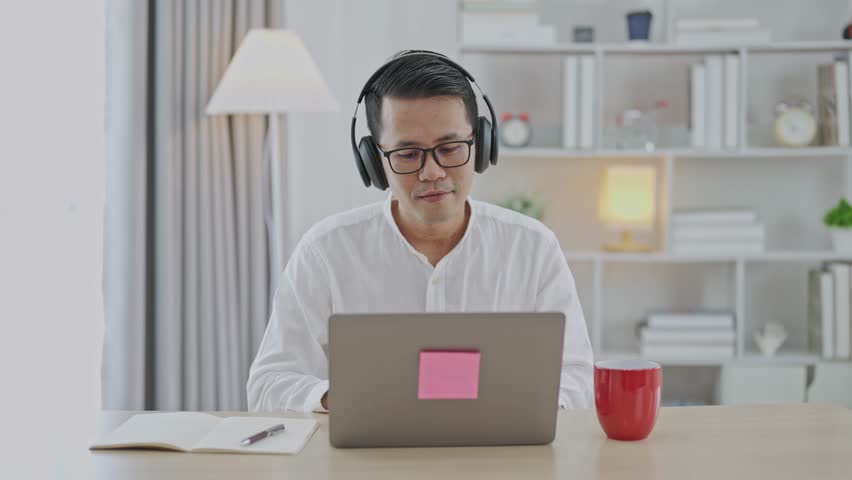 Asian freelance business man wearing headphones making video call in home office, using laptop, talking, watching webinar or studying language, speaking, online training, explaining, e-learning. Royalty-Free Stock Footage #1111765717