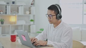 Asian freelance business man wearing headphones making video call in home office, using laptop, talking, watching webinar or studying language, speaking, online training, explaining, e-learning.