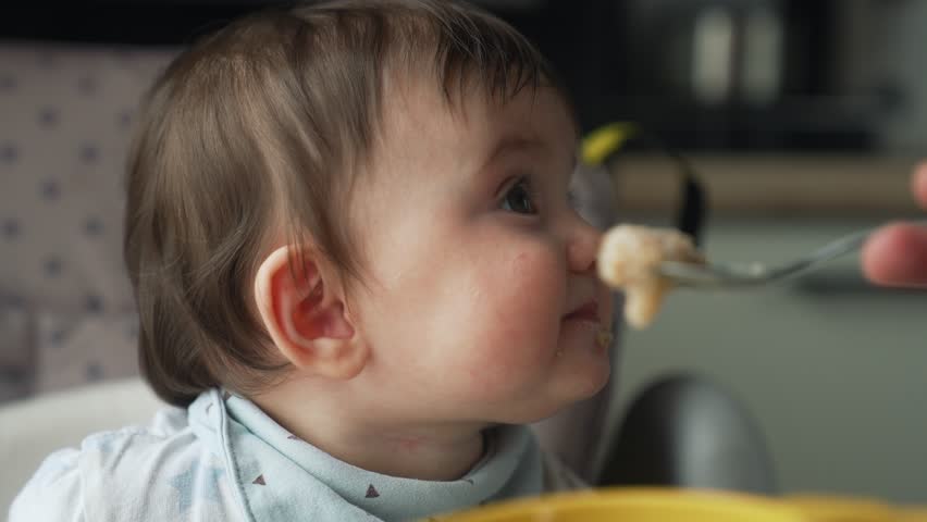 Happy family concept. close-up portrait of a baby sitting on chair and eating from a spoon. introduction of baby food in kindergarten.nanny feeds baby healthy food from box. breakfast in kindergarten Royalty-Free Stock Footage #1111766507