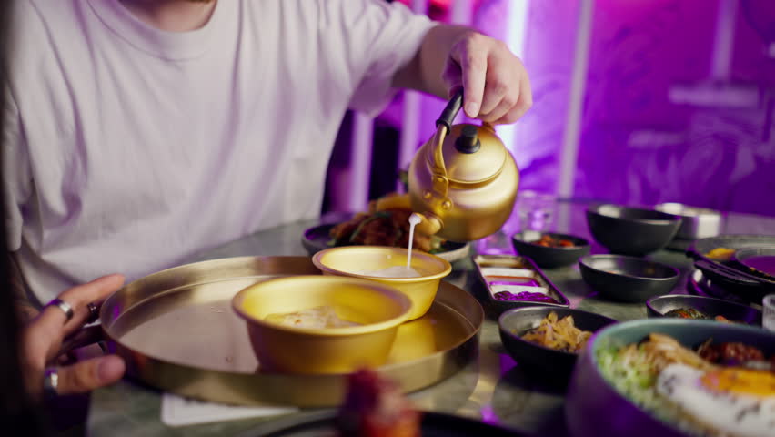 close-up of a girl pouring a traditional Korean low-alcohol drink from a golden teapot into cup with ice in restaurant