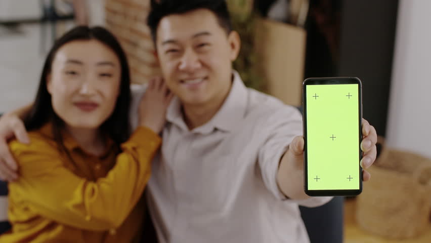 Best family app. Blurred asian couple bonding at home and showing smartphone with green chroma key screen and tracking marks in male hand, selective focus, slow motion, empty space | Shutterstock HD Video #1111772433