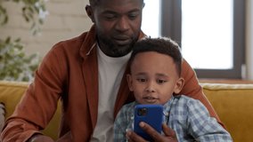 Learning together. Close up portrait of bonding african american father and son watching video on smartphone and discussing it, resting at home, tracking shot