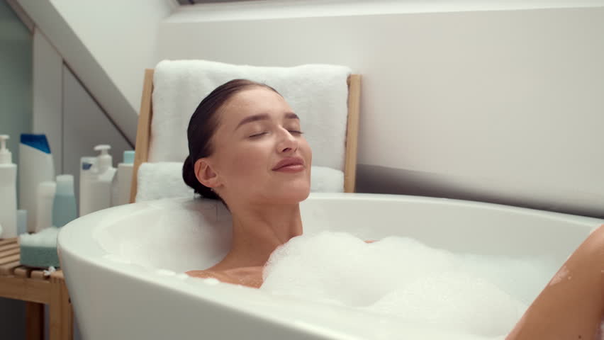 Slow motion 4K video footage showcases a happy and delighted woman, deeply immersed in a luxurious foamy bath. The scene exudes tranquility, relaxation, and the pure joy of self-pampering moments. | Shutterstock HD Video #1111772445