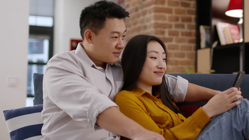 Gadget lifestyle concept. Chinese married couple using smartphone, relaxing on weekend, bonding on couch at home, spending time together, tracking shot, slow motion | Shutterstock HD Video #1111772447