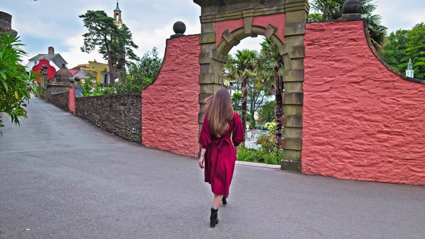 A beautiful girl in a red dress visits Portmeirion village. Female tourist looking at the beautiful coloured houses and garden in Wales.