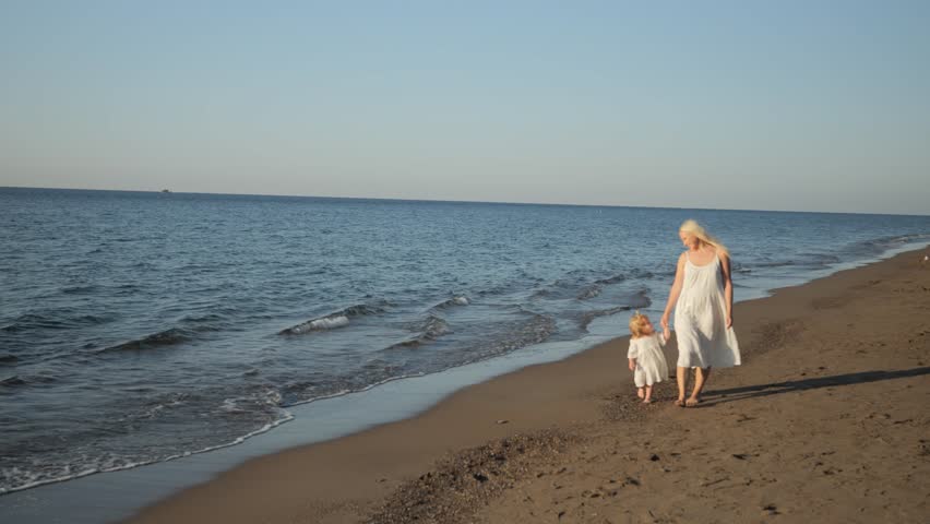 Mom and daughter on a walk during a vacation at the sea are walking along the beach dressed in white dresses and holding hands. A happy young mother and her daughter. High quality 4k footage | Shutterstock HD Video #1111775393