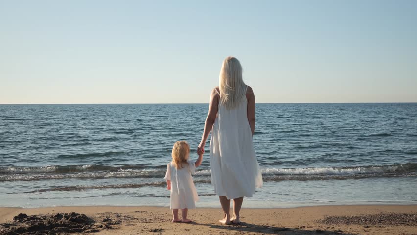 Happy childhood of young children, a young mother and daughter of 2s are standing on the beach near the water and looking at the blue sea waves. Vacation of a mother and her beloved daughter | Shutterstock HD Video #1111775395