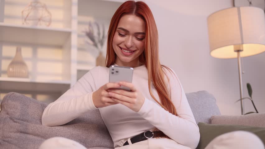 Charming young red haired woman hold smartphone scrolling app watching social media feed and looking at the screen indoors Happy female texting enjoying leisure time at cozy home | Shutterstock HD Video #1111778953