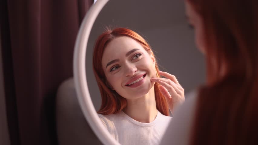 Portrait of pretty young red haired woman touching facial skin check quality of care products while looking at mirror reflection at home Healthy skin care treatment beauty concept | Shutterstock HD Video #1111778957
