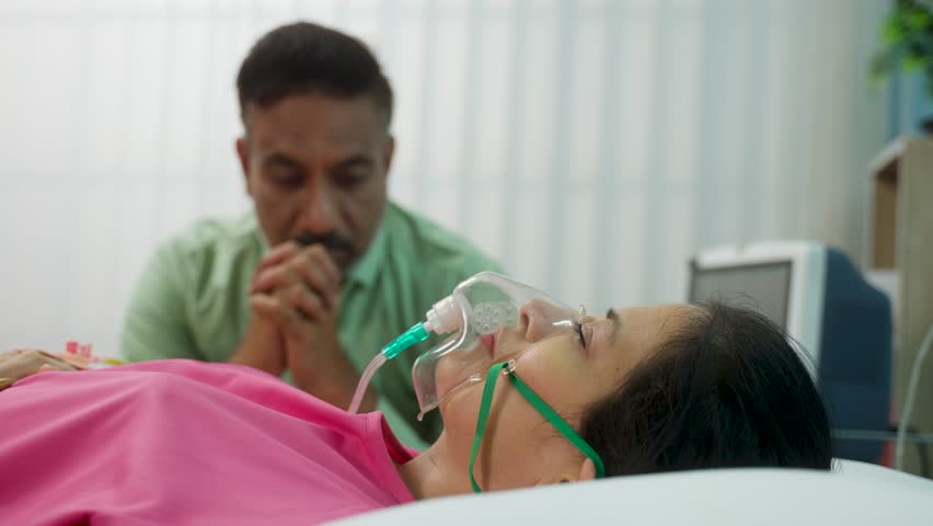 Rack focus shot, Indian senior husband praying for medical recovery of sick wife breathing on oxygen ventilator at hospital - concept of health care emergency, family support and compassion | Shutterstock HD Video #1111781087