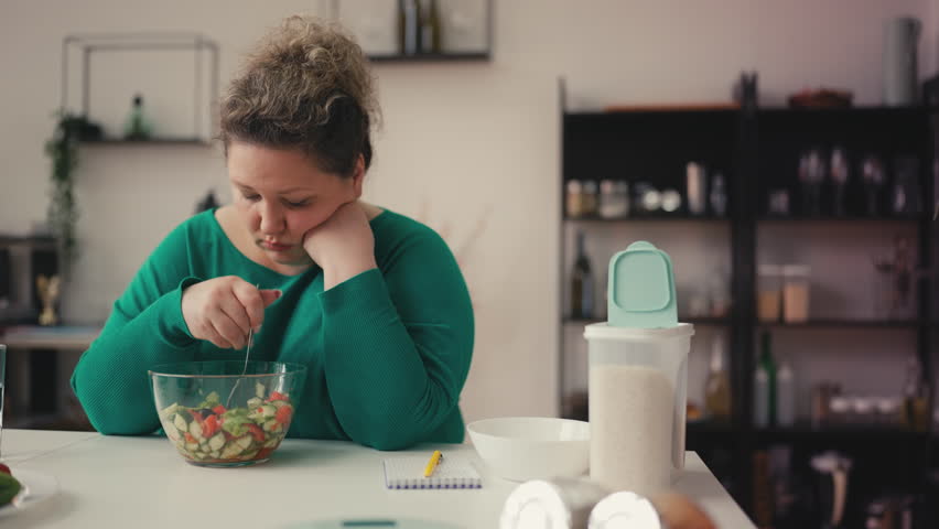 Sad overweight woman eating vegetable salad with unhappy face, weightloss diet