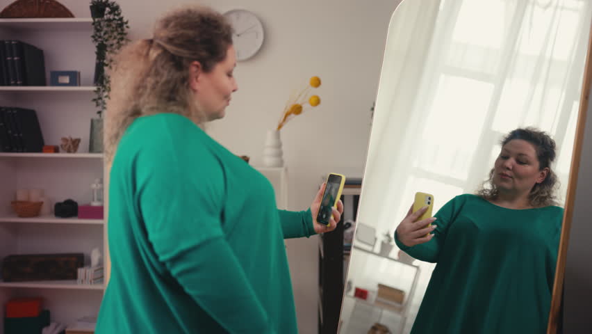Confident body positive woman taking selfie on smartphone, posing near mirror